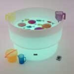 Mesa de experimentación con agua, arena y luz led encendida en color blanco, rellena de agua y con jarras para trasvases.