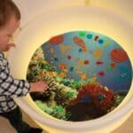 Niño jugando con las conchas translúcidas de colores en un minimundo marino montado en la mesa sensorial de agua y luz.