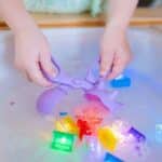 Manos utilizando la pala recogedor incluida en el pack de Niño jugando con el cubo tamizador del pack de Herramientas sensoriales Pick up Pals