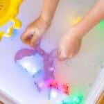 Manos utilizando la pala recogedor incluida en el pack de Niño jugando con el cubo tamizador del pack de Herramientas sensoriales Pick up Pals