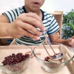 Niño haciendo trasvases sólidos con alubias y un cazo en una bandeja de actividad de la vida práctica montessori. Actividad incluida en el imprible Guía de actividades para la vida practica montessori